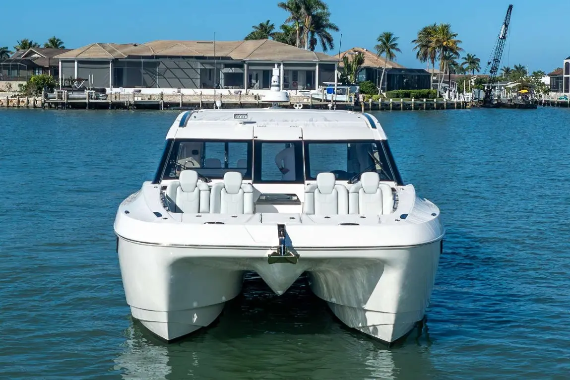 A sleek white boat with cushioned seats floats on clear blue water, near waterfront homes and palm trees.